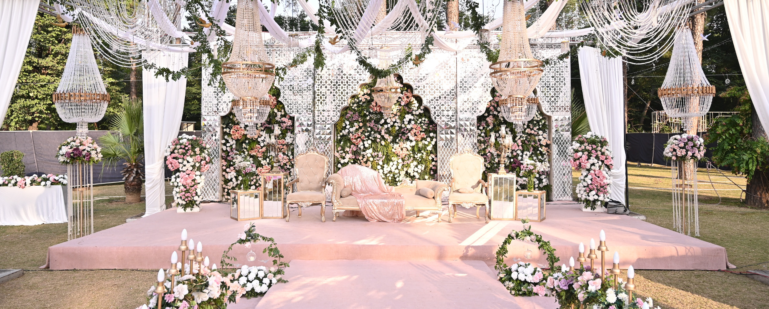 Opulent pink and white floral wedding reception stage with elaborate lattice backdrop, large crystal chandeliers, and white tufted chairs.