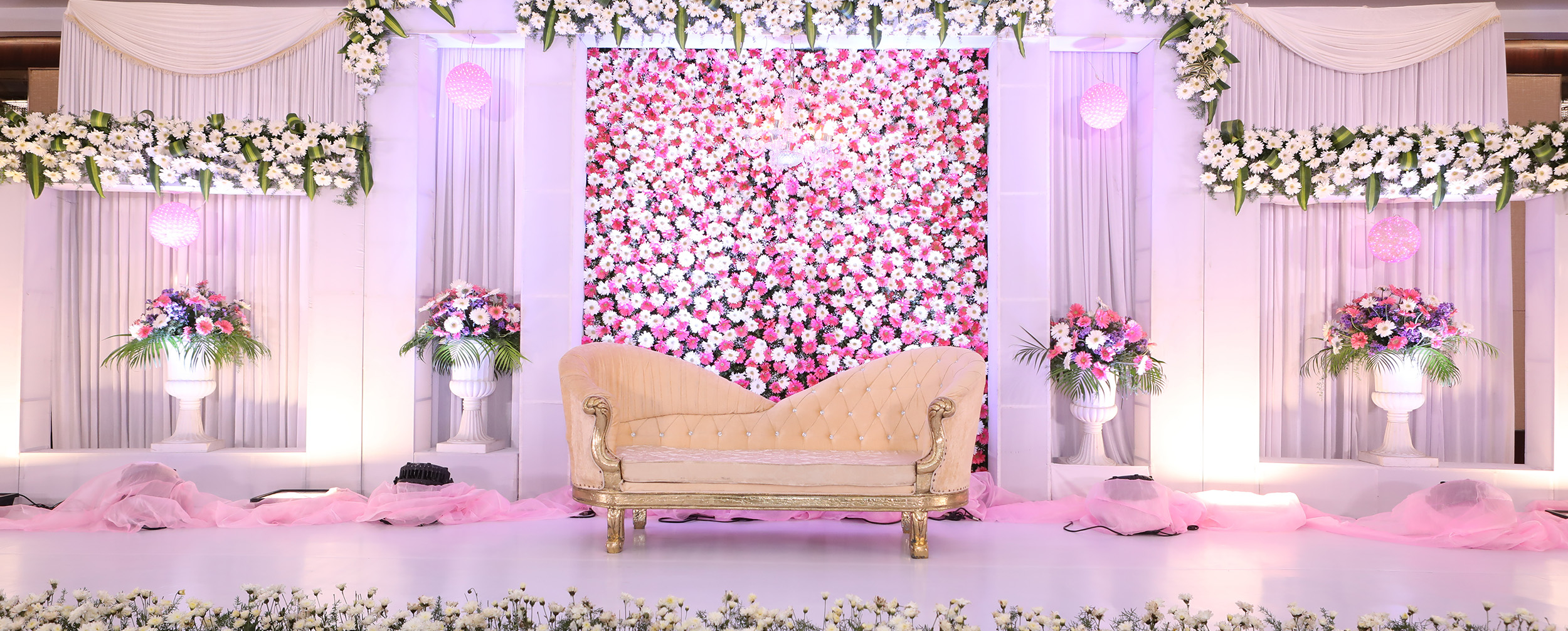 Elegant wedding reception stage featuring a flower wall backdrop of pink and white roses and a gold-framed chaise lounge.